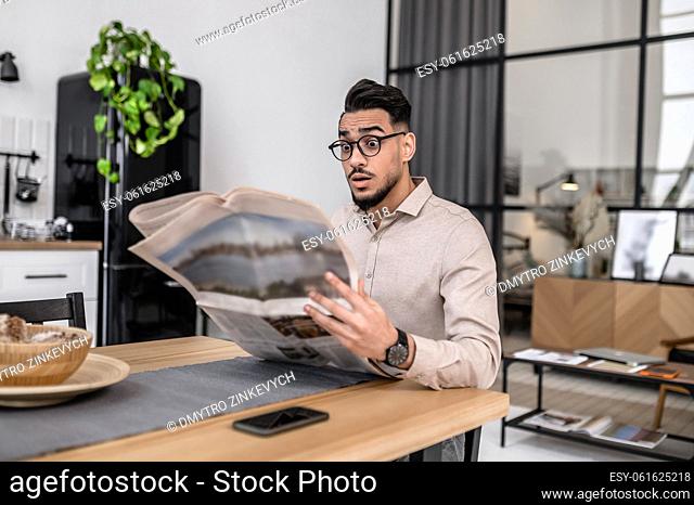 Surprise, news. Surprised young bearded man in glasses with open mouth reading newspaper sitting at table at home
