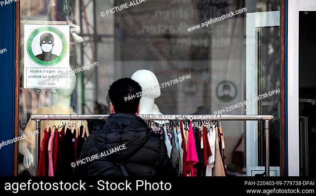 03 April 2022, North Rhine-Westphalia, Duesseldorf: A woman looks at clothes on a rack outside a store, a sign in the window reads ""Wear mask to protect others