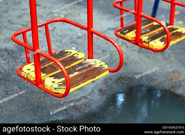 Red metal swing with a wooden seat on a kids playground on a rainy day with a puddle under it