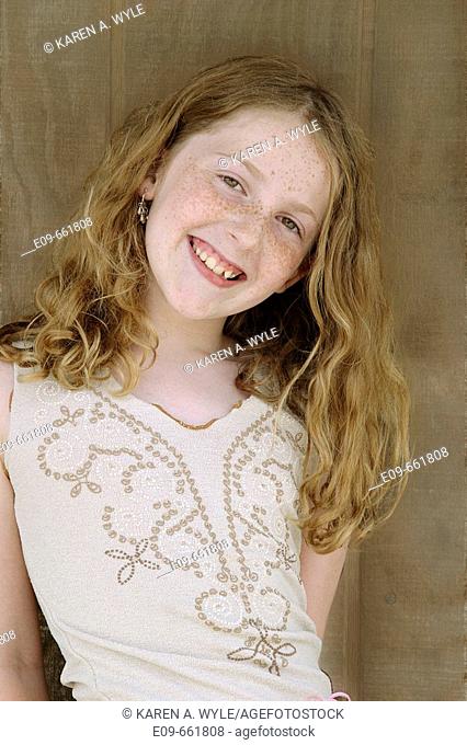 Preteen girl with long gold-brown hair and freckles, smiling at camera, head tilted to side, waist up, against brown wood background