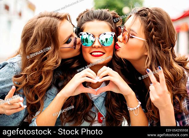 Portrait of beautiful ladies with hairstyles and make up wearing stylish sunglasses and showing peace signs while kissing friend in mirrored sunglasses with red...