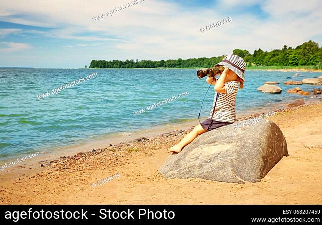 Little girl sitting on the stone on the seashore with binocular in her hands. Concept of the childhood and family vacation