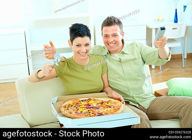 Happy couple sitting on sofa in, living room, eating pizza, smiling