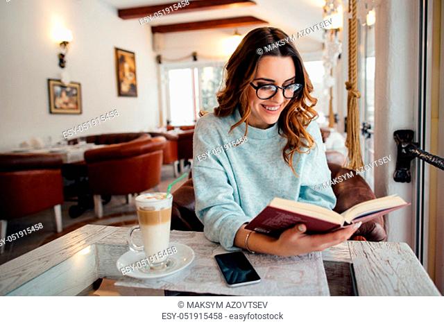Young beautiful woman in glasses reading interesting book in cafe