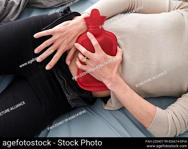 PRODUCTION - 30 March 2022, Berlin: ILLUSTRATION - A woman holds a hot water bottle to her lower abdomen while lying in bed