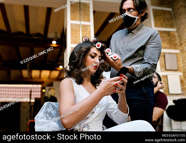 Male hairdresser doing hairstyle while bride using smart phone at salon during pandemic