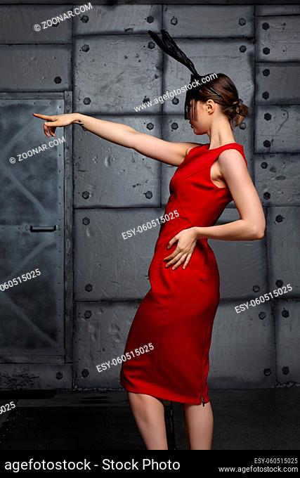 Young woman in black rabbit or hare fancy mask and red dress. Arm is stretched out. In the background there is a metal wall with rivets