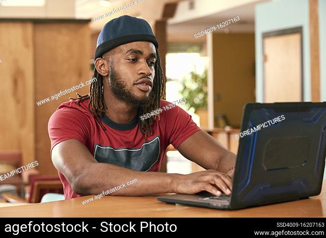 Portrait of young ethnic man wearing red t shirt and knit hat , sitting at bar in kitchen of downtown loft with laptop computer