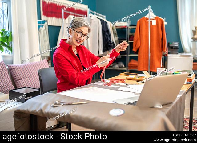 Interest. Stylish woman holding measure looking inquiringly at laptop sitting at table with pattern and sewing accessories in atelier