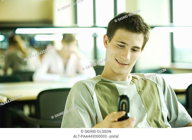 Male college student sitting in study room, looking at cell phone, smiling