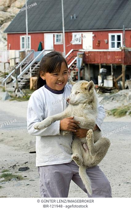 Young Inuk girl with a greenland dog puppy with typical houses in background, Umanak, Greenland