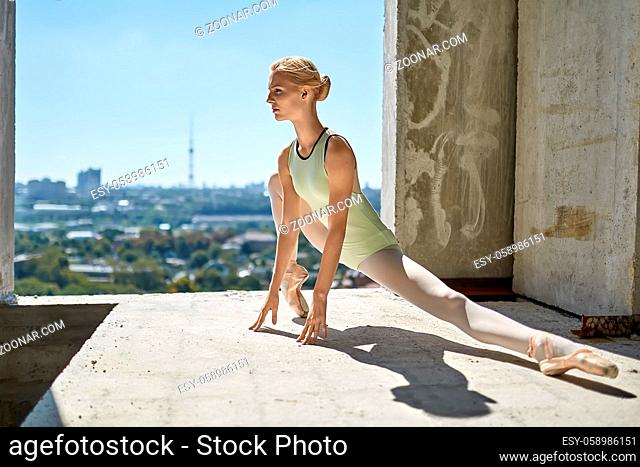 Delightful ballerina is posing on the concrete floor of the unfinished building on the cityscape background. She wears a green leotard with light leggings and...