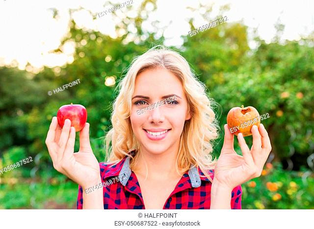 Beautiful young blond woman in checked red shirt harvesting apples, holding in her hands one rotten and one good apple