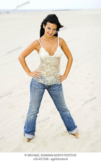 A 20's 30's young woman wearing blue jeans and a lace top, stands on beach barefoot with hands on her hips