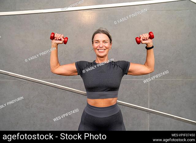 Smiling mature woman exercising with dumbbells in front of wall