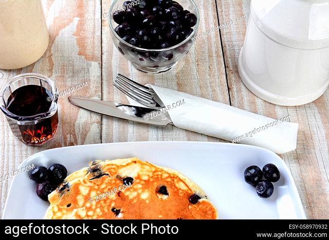 Closeup of a stack of fresh homemade blueberry pancake breakfast on white plate. Maple Syrup utensils and corckery and a rustic wood table
