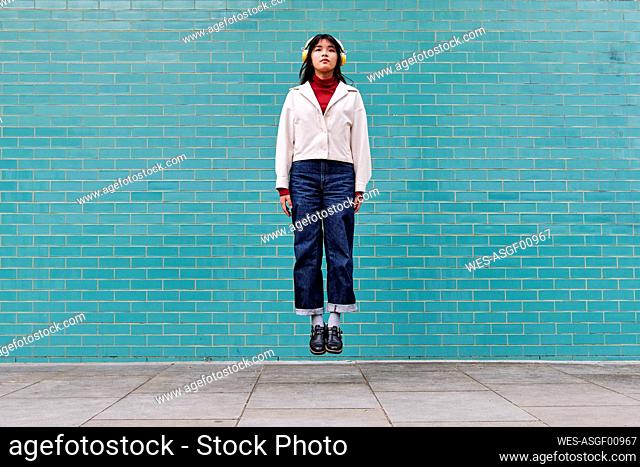 Woman with headphones levitating in front of turquoise brick wall
