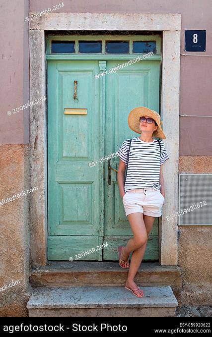 Beautiful young female tourist woman wearing sun hat, standing and relaxing in shade in front of vinatage wooden door in old Mediterranean town while...