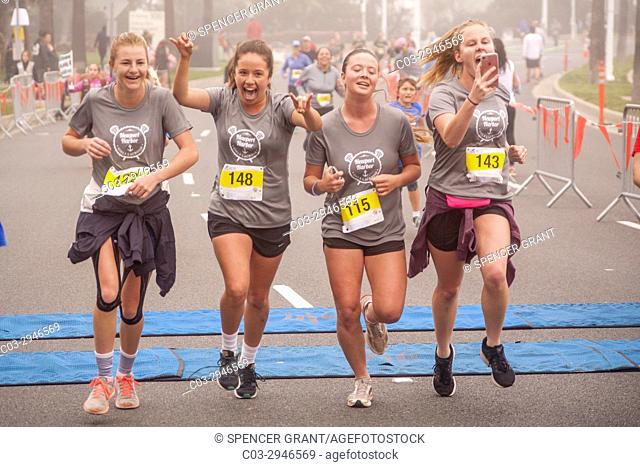 Four exuberant teen girl runners cross the finish line at a community foot race in Newport Beach, CA. Note one checking running time on her cell phone
