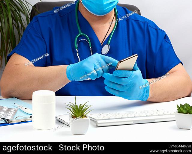 doctor in a blue uniform sits at a desk and holds in his hand a smartphone, concept of distance medicine and consultations