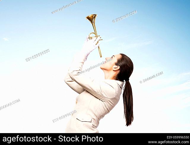 Beautiful woman playing trumpet brass. Young businesslady in white business suit and gloves posing with music instrument on blue sky background