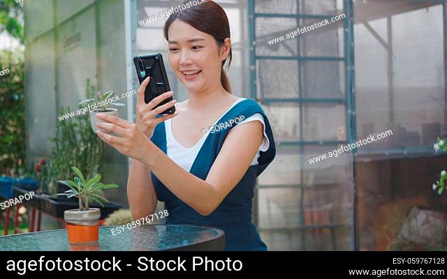 Asian beautiful young woman taking photos of small tree on pot by smartphone for sale in garden near greenhouse, female gardener photography flowers on digital...
