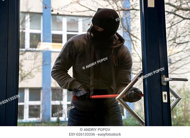 Full length of hooded man using crowbar to open glass door