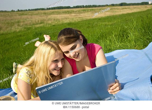Two smiling girls using a laptop while lying on a blanket on a meadow