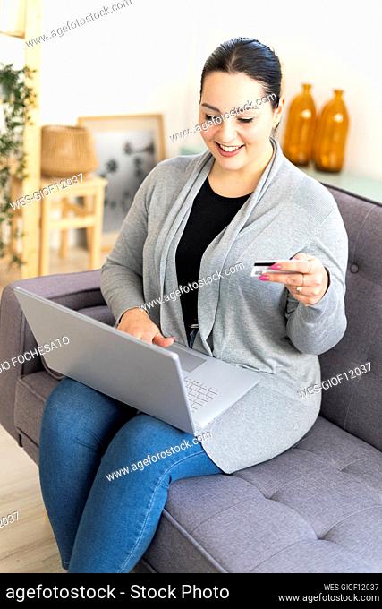 Smiling woman holding credit card while shopping online using laptop at home
