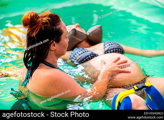 Female therapist giving belly water massage to pregnant woman in bikini with rubber neck pillow around her neck and leg. support float in swimming pool