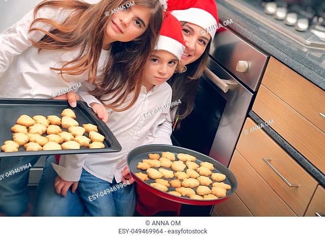 Happy mother with two cute kids baking tasty sweet homemade cookies, gingerbread, traditional Christmas sweets, good Christmastime family tradition