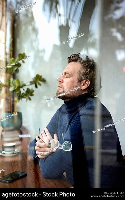 Businessman with eyes closed sitting in coffee shop seen through glass window