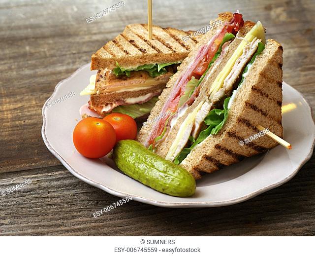 Photo of a club sandwich made with turkey, bacon, ham, tomato, cheese, lettuce, and garnished with a pickle and two cherry tomatoes