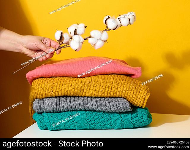 stack of folded knitted sweaters and a woman's hand holding a branch of cotton on a yellow background