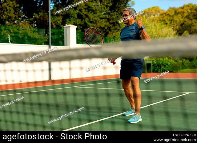 Cheerful biracial senior man holding racket gesturing while playing tennis in court seen through net