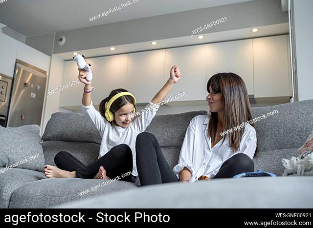 Cheerful daughter with hand raised sitting by mother while playing video games in living room