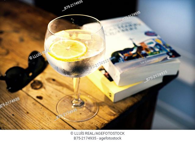 Closeup of a glass of gin and tonic on a table with some books and sunglasses