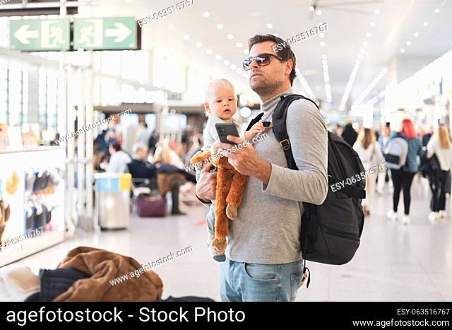 Father traveling with child, holding his infant baby boy at airport terminal, checking flight schedule, waiting to board a plane