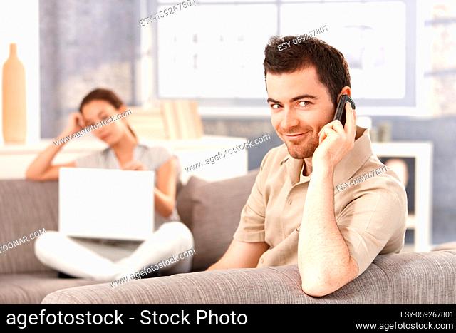 Young man sitting on sofa at home, talking on mobile, smiling, woman using laptop in the background