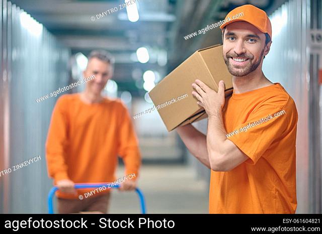 Joyous young loader holding the cardboard box on his shoulder standing beside his male colleague