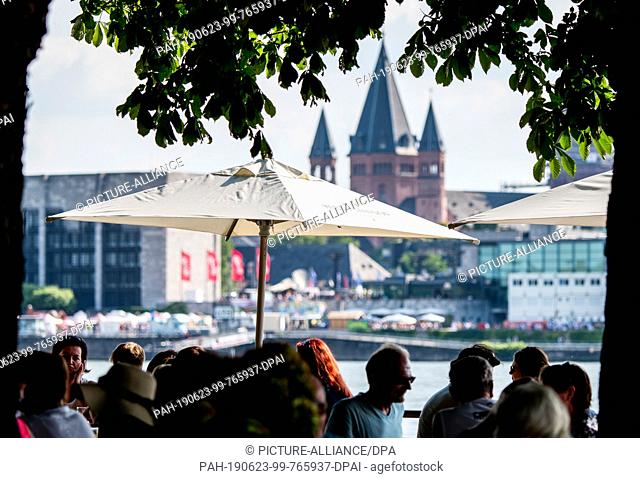 23 June 2019, Hessen, Wiesbaden: People sit in Mainz-Kastel under umbrellas and chestnut trees on the banks of the Rhine with a view of Mainz Cathedral