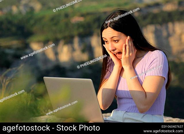 Excited asian woman watching laptop media content sitting in nature