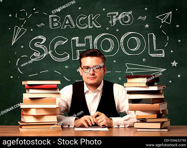 A young teacher in glasses sitting at classroom desk with pile of books in front of blackboard saying back to school drawing concept