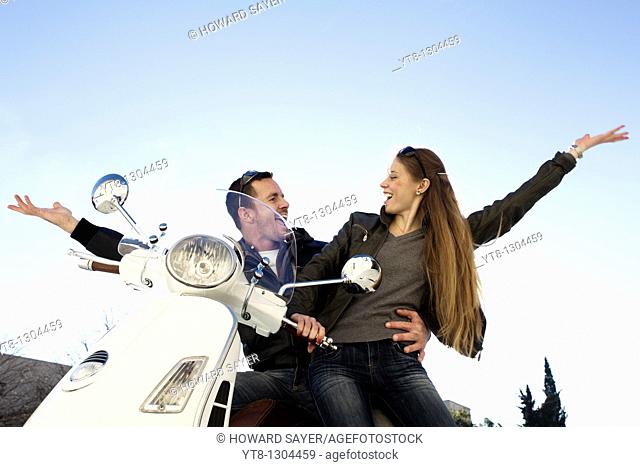Couple sitting on a motor scooter and raising their arms in the air