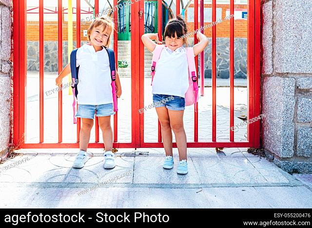 A black-haired girl wearing a pink backpack and a blonde-haired girl wearing a blue backpack at the school entrance door. School concept