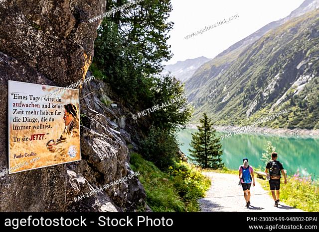 24 August 2023, Austria, Brandberg: A woman and a man walk along a hiking trail at the Zillergrund reservoir past richly overgrown mountain slopes