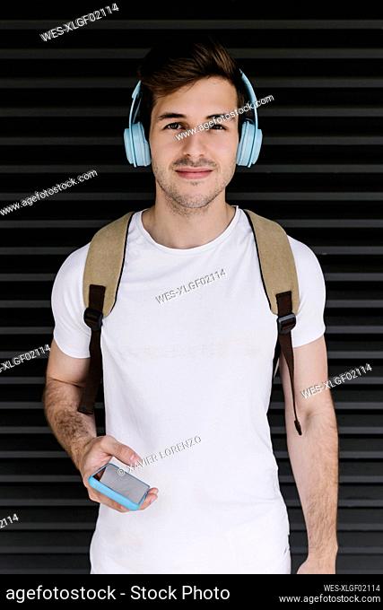 Young man holding mobile phone while listening music through headphones in front of shutter