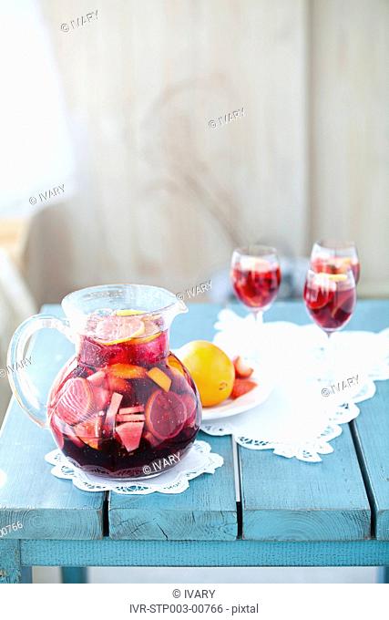 Jug full of red wine mixed with fruits with glasses in background
