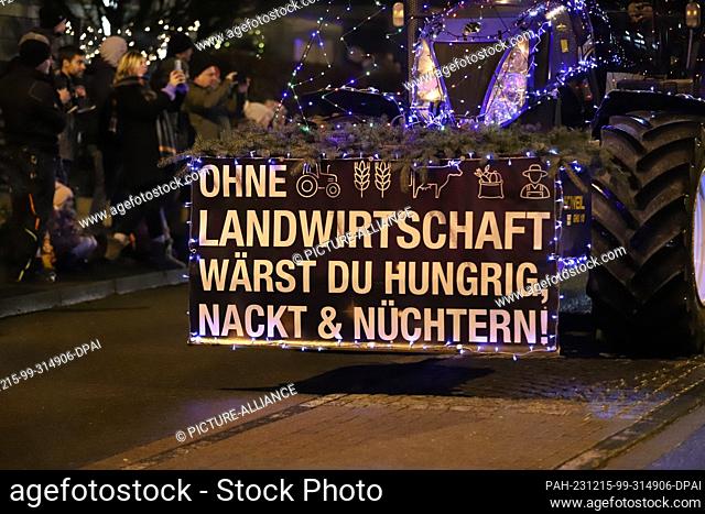 15 December 2023, North Rhine-Westphalia, Kürten: A sign reading ""Without agriculture you would be hungry, naked & sober"" is attached to a lighted tractor...