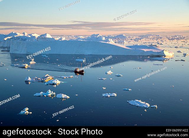 Ilulissat, Greenland - July 7, 2018: A red fishing boat sailing in between icebergs on Ilulissat Icefjord